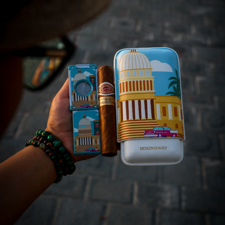 Hand holding two cigars with decorative boxes featuring architectural designs on a textured surface.